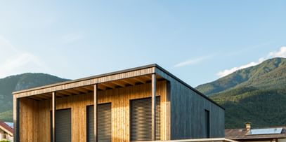 Modernes zweistöckiges Haus mit großen Fenstern und Holzfassade, in der Abenddämmerung beleuchtet. Das Haus ist von grünem Rasen und einem gepflegten Garten umgeben, im Hintergrund sind Berge und weitere Häuser zu sehen.