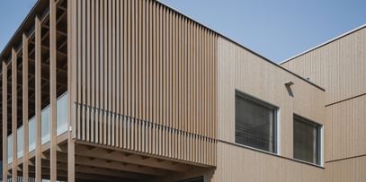 Gebäude mit Holzfassade und großen Fenstern. Eine offene Treppe führt zu einem erhöhten Eingangsbereich.