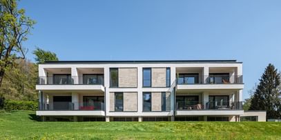 Modernes zweistöckiges Gebäude mit großen Fenstern und Balkonen, gelegen in einer grünen Umgebung mit Bäumen im Hintergrund bei sonnigem Wetter und klarem Himmel.