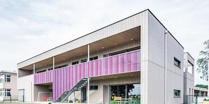 Zweigeschossiges Gebäude, deren Außenwände aus hellem Holz gefertigt sind und ein markanter, lila Streifen die obere Etage ziert