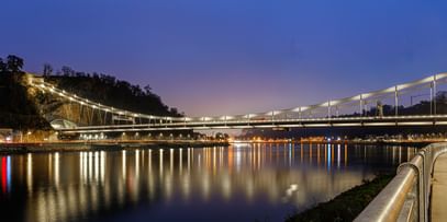 Eine nächtliche Luftaufnahme einer Brücke, die über einen Fluss führt. Die Brücke ist beleuchtet und reflektiert im Wasser.