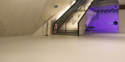 Moderner Ausstellungsraum mit weißen Wänden und Bodenbeschichtung im Schlossmuseum Linz. eine Treppe mit schwarzem Glasgeländer führt hinauf zum Ausgang