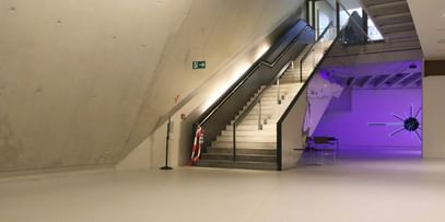 Moderner Ausstellungsraum mit weißen Wänden und Bodenbeschichtung im Schlossmuseum Linz. eine Treppe mit schwarzem Glasgeländer führt hinauf zum Ausgang