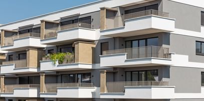 Wohnanlage in der Robert-Stolz-Straße in Linz. Ein modernes, mehrstöckiges Wohngebäude mit hellen Fassaden und zahlreichen Balkonen. Große Fenster sorgen für viel Tageslicht. Vor dem Gebäude stehen Autos auf einer ruhig wirkenden Straße, eingerahmt von gepflegten Grünflächen und Bäumen.