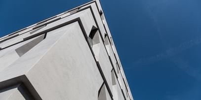 Die Ecke eines Gebäudes, die in die Höhe ragt. Die Fassade ist aus hellem Beton mit schmalen Fenstern, vor einem klaren blauen Himmel.