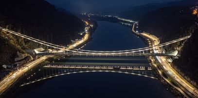 Eine nächtliche Luftaufnahme einer Brücke, die über einen Fluss führt. Die Brücke ist beleuchtet und reflektiert im Wasser.