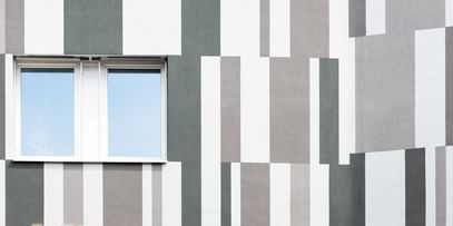 Ein Gebäude mit einer Fassade, die aus vertikalen und horizontalen Streifen in verschiedenen Grautönen und Weiß besteht. In der Mitte der Wand befindet sich ein Fenster mit zwei Fensterscheiben.
