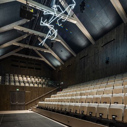 Auditorium mit einer Holzdecke und einer Sitzanordnung aus hellen, gepolsterten Stühlen. An der Decke hängt eine künstlerische Neoninstallation. Die Wände sind aus dunklem Holz, und es gibt eine Bühne im Vordergrund.