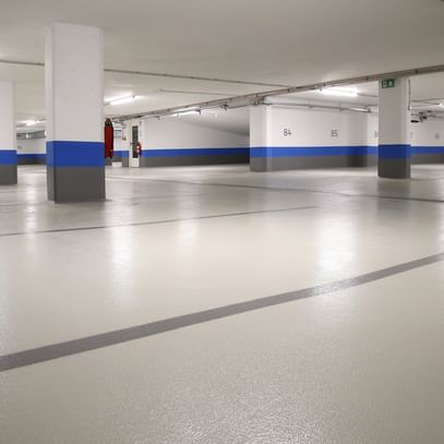 Ein leerer Parkhausbereich mit hellen Wänden und einem grauen Boden. Die Wände sind teilweise mit blauen Streifen dekoriert. Es gibt mehrere Säulen und einige Feuerlöscher an den Wänden.