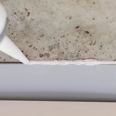Ein Silikon- oder Dichtmittel wird aus einer Tube auf eine Fuge zwischen einem Betonuntergrund und einer weißen Wandverkleidung aufgetragen.