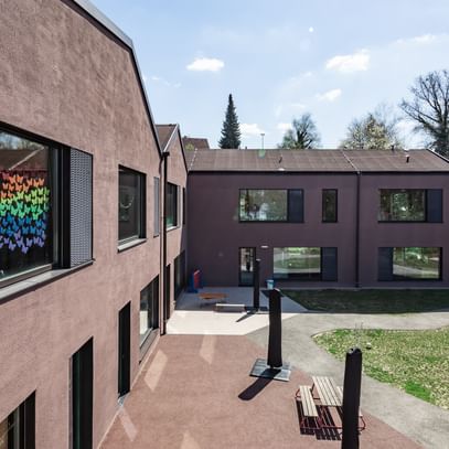 Zweigeschossiges Gebäude mit großen Fenstern und schwarzen Fensterläden. In den oberen Fenstern sind bunte Zeichnungen sichtbar.