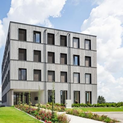 Ein mehrstöckiges Gebäude mit einer Fassade aus Holz und großen Fenstern. Es hat eine rechteckige Form und ist von einem gepflegten Garten mit Blumen und Sträuchern umgeben