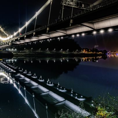 Eine nächtliche Luftaufnahme einer Brücke, die über einen Fluss führt. Die Brücke ist beleuchtet und reflektiert im Wasser.