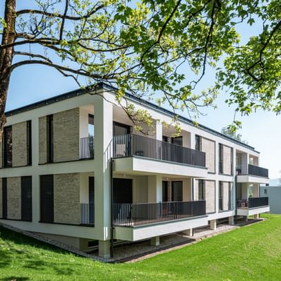 Modernes zweistöckiges Gebäude mit großen Fenstern und Balkonen, gelegen in einer grünen Umgebung mit Bäumen im Hintergrund bei sonnigem Wetter und klarem Himmel.