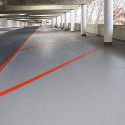 Der Boden einer Parkgarage ist in einem hellen Grauton gestrichen, während orangefarbene Linien diagonal über den Boden verlaufen, um Parkplätze oder Verkehrswege zu kennzeichnen.