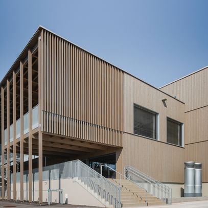 Gebäude mit Holzfassade und großen Fenstern. Eine offene Treppe führt zu einem erhöhten Eingangsbereich.
