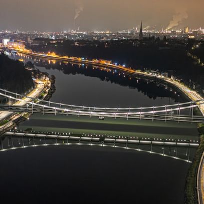 Eine nächtliche Luftaufnahme einer Brücke, die über einen Fluss führt. Die Brücke ist beleuchtet und reflektiert im Wasser.