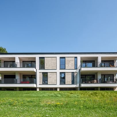 Modernes zweistöckiges Gebäude mit großen Fenstern und Balkonen, gelegen in einer grünen Umgebung mit Bäumen im Hintergrund bei sonnigem Wetter und klarem Himmel.