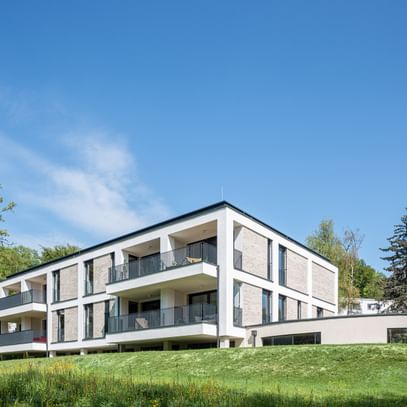 Modernes zweistöckiges Gebäude mit großen Fenstern und Balkonen, gelegen in einer grünen Umgebung mit Bäumen im Hintergrund bei sonnigem Wetter und klarem Himmel.
