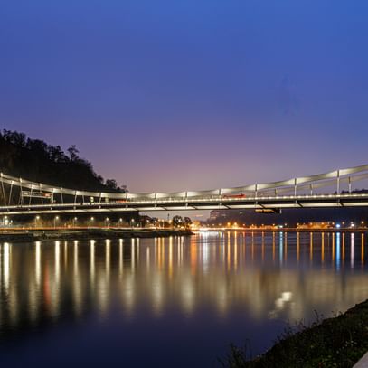 Eine nächtliche Luftaufnahme einer Brücke, die über einen Fluss führt. Die Brücke ist beleuchtet und reflektiert im Wasser.