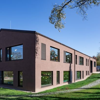 Zweigeschossiges Gebäude mit großen Fenstern, das im Grünen steht. In den Fenstern sind bunte Zeichnungen sichtbar.