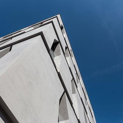 Die Ecke eines Gebäudes, die in die Höhe ragt. Die Fassade ist aus hellem Beton mit schmalen Fenstern, vor einem klaren blauen Himmel.