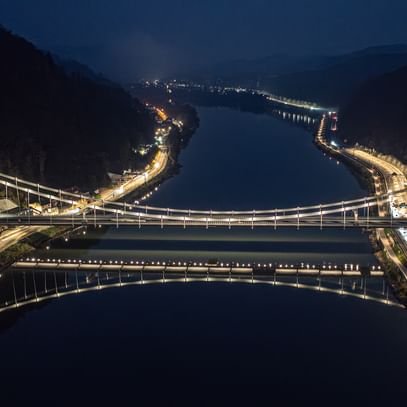 Eine nächtliche Luftaufnahme einer Brücke, die über einen Fluss führt. Die Brücke ist beleuchtet und reflektiert im Wasser.