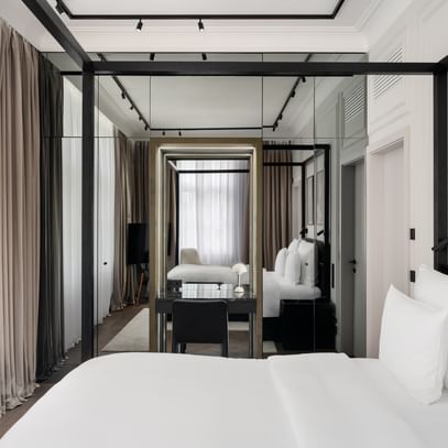 Helles Executive-Suite-Zimmer im „The Amauris“ Wien mit großem Doppelbett, weißer Bettwäsche, Spiegelfront mit Schreibtisch und schwarzem Stuhl, bodentiefen Vorhängen und gemütlicher Sitzecke mit Sessel und TV im Hintergrund.