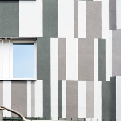 Ein Gebäude mit einer Fassade, die aus vertikalen und horizontalen Streifen in verschiedenen Grautönen und Weiß besteht. In der Mitte der Wand befindet sich ein Fenster mit zwei Fensterscheiben.