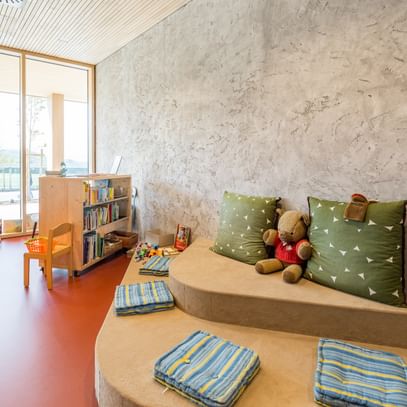 Im Vordergrund befinden sich zwei große, grüne Kissen auf einer erhöhten Sitzfläche aus hellem Holz. Daneben liegen gestreifte Decken. Im Hintergrund steht ein Bücherregal mit einer Auswahl an Kinderbüchern. Auf dem Boden sind einige Spielzeuge und ein Teddybär zu sehen.