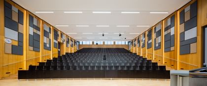 Leerer Hörsaal mit gestuften Sitzreihen, einem Rednerpult mit elektronischer Ausstattung und dekorativen Akustikpaneelen in verschiedenen Formen und Farben an den Holzverkleidungen.