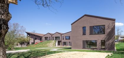 Zweigeschossiges Gebäude mit brauner Fassade und großen Fenstern, umgeben von einer grünen Wiese und Bäumen. Im Vordergrund ist ein Sandbereich sichtbar.