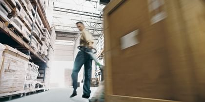 Ein Arbeiter in einem Lager zieht einen großen Holzbehälter auf einem Rollwagen. Im Hintergrund sind Regale mit weiteren Materialien und Kisten zu sehen.