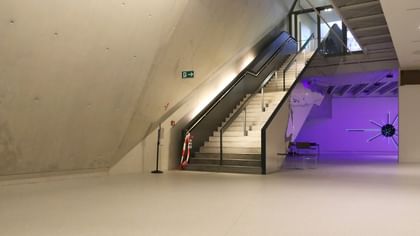 Moderner Ausstellungsraum mit weißen Wänden und Bodenbeschichtung im Schlossmuseum Linz. eine Treppe mit schwarzem Glasgeländer führt hinauf zum Ausgang