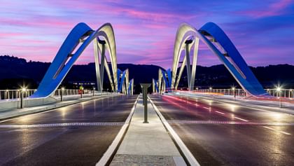 Brücke mit geschwungenen, weißen Bögen, die sich über eine Straße erstreckt. Der Himmel ist in sanften Lila- und Rosatönen gefärbt.