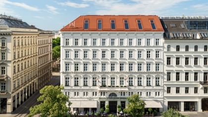 Außenansicht des „The Amauris“ Hotels in Wien mit klassizistischer weißer Fassade, symmetrischen Fenstern, rotem Mansarddach, Straßenbäumen im Vordergrund und blauem Himmel.