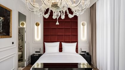 Schlafzimmer der Opera Suite im „The Amauris“ Wien mit großem Doppelbett vor hohem, weinrotem Kopfteil, vertikalen Lichtleisten in weißen Wandpanelen, opulentem Glaslüster und Einblick in ein marmorverkleidetes Badezimmer.