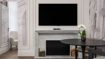 Wohnbereich einer Suite im „The Amauris“ Wien mit Kaminfront aus hellem Marmor, darüber ein Flachbildfernseher, runder Tisch mit zwei Stühlen und weißer Orchidee sowie Blick in einen angrenzenden Raum mit Sessel und bodenlangen Vorhängen auf dunklem Fischgrätparkett.