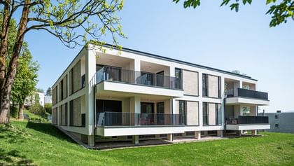 Modernes zweistöckiges Gebäude mit großen Fenstern und Balkonen, gelegen in einer grünen Umgebung mit Bäumen im Hintergrund bei sonnigem Wetter und klarem Himmel.
