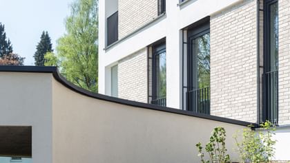 Modernes Gebäude mit weißer und heller Ziegelfassade, großen Fenstern mit schwarzen Rahmen und einem überhängenden Bauteil. Im Vordergrund ein bepflanzter Hang, im Hintergrund Bäume und blauer Himmel.