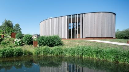 Rundes Gebäude mit einer Holzfassade, das auf einem sanften Hügel steht. Im Vordergrund ist ein ruhiger Teich zu sehen