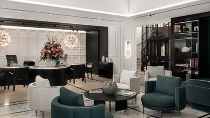 Elegante Lobby im „The Amauris“ Wien mit dunkelgrünen und hellgrauen Lounge-Sesseln um schwarze Couchtische, geschwungenem Empfangstresen mit Blumenarrangement, zwei kugelförmigen Designerleuchten, Marmortreppe neben historischem Aufzug und Regalen sowie weißen Wandpanelen mit indirekter Beleuchtung.