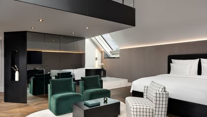 Großzügige Grand-Maisonette-Suite im „The Amauris“ Wien mit Kingsize-Bett, gepolstertem grauen Kopfteil, smaragdgrünen Lounge-Sesseln, kariertem Sessel, schwarzem Couchtisch, Spiegelwand und Galerieebene unter einer Dachschräge mit Oberlicht.