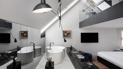 Loftartige Grand-Maisonette-Suite im „The Amauris“ Wien mit zwei freistehenden runden Badewannen auf hellem Marmorboden, langen schwarzen Marmorablagen mit Deko, großem Doppelbett und Galerieebene mit Glasbrüstung unter schräger Dachdecke.