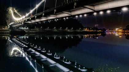 Eine nächtliche Luftaufnahme einer Brücke, die über einen Fluss führt. Die Brücke ist beleuchtet und reflektiert im Wasser.