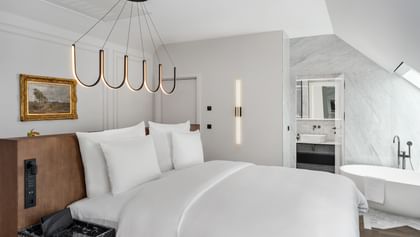 Modernes Schlafzimmer der Loft Suite im „The Amauris“ Wien mit großem Doppelbett, Holz-Kopfteil, organischer schwarzer Deckenleuchte, Wandgemälde und Blick in ein helles Badezimmer mit freistehender Badewanne und Marmorflächen.