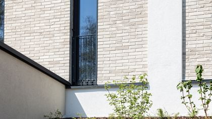  Nahaufnahme einer Gebäudefassade mit hellen Ziegeln, schwarzen Fensterbänken und Metallgittern an den Fenstern. Unten und rechts ist weißer Putz zu sehen, entlang der Wand verläuft ein Streifen mit weißen Kieselsteinen.