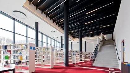 Moderner, heller Bibliotheksraum der Wirtschaftsuniversität Wien mit rotem Teppich, weißen Bücherregalen, großen Glasfronten und schwarzen Säulen. Eine breite Treppe führt ins offene, lichtdurchflutete Obergeschoss.