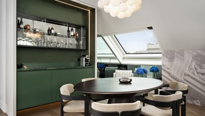 Ess- und Loungebereich der Loft Suite im „The Amauris“ Wien mit rundem Holztisch und vier gepolsterten Stühlen, eleganter grüner Einbau-Bar mit Spiegelregal, Flaschen und Gläsern sowie großen Dachfenstern mit Stadtblick und blauen Hortensien.