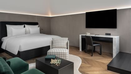 Eleganter Wohn- und Schlafbereich in der Grand-Maisonette-Suite des „The Amauris“ Wien mit großem Doppelbett, Schreibtisch mit TV, zwei dunkelgrünen Sesseln, schwarzem Couchtisch mit Schachbrett und kariertem Sessel auf hellem Teppich und Fischgrätparkett.